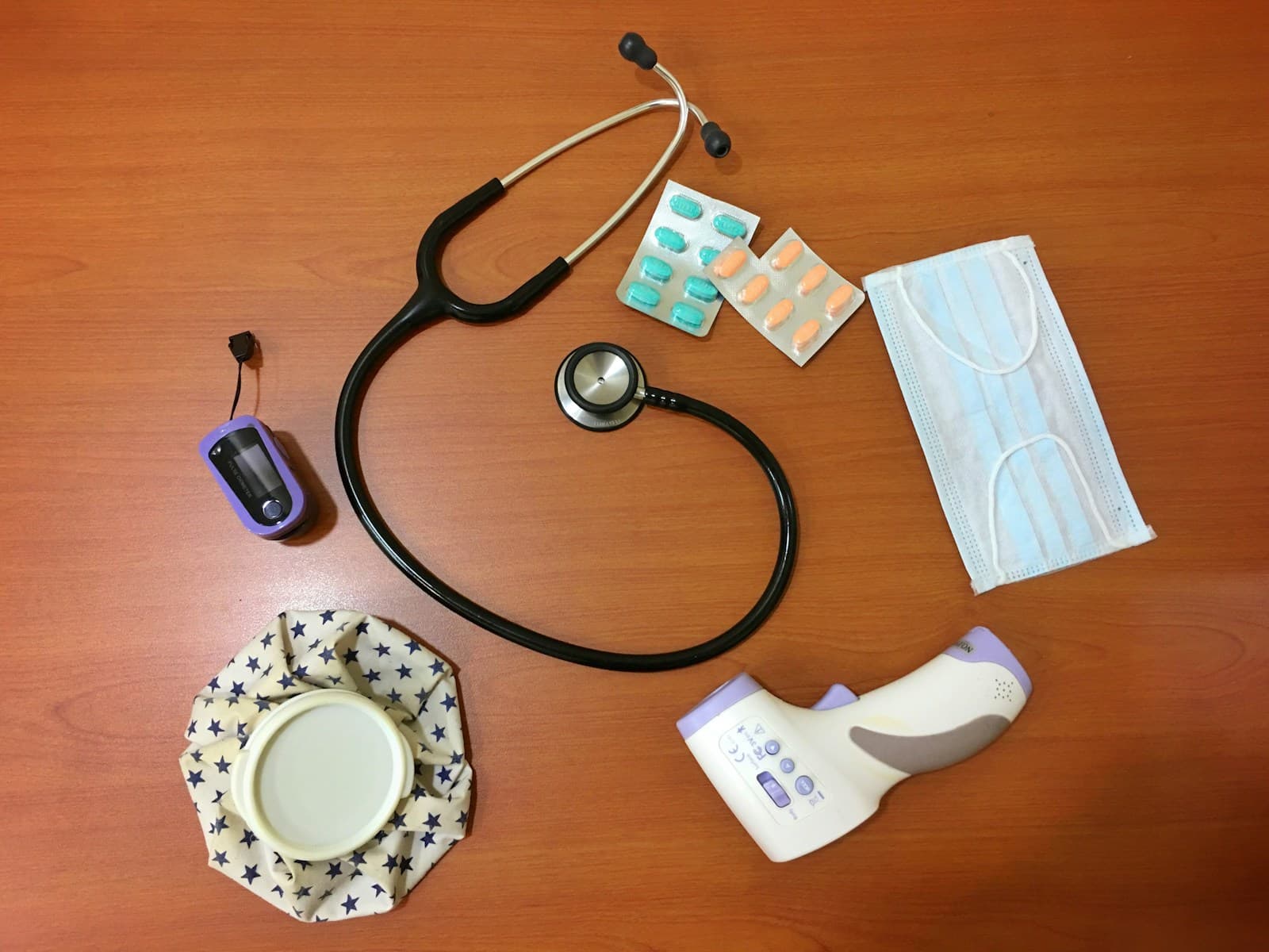 A stethoscope, pulse oximeter, mask and other medical supplies on a wooden desk.