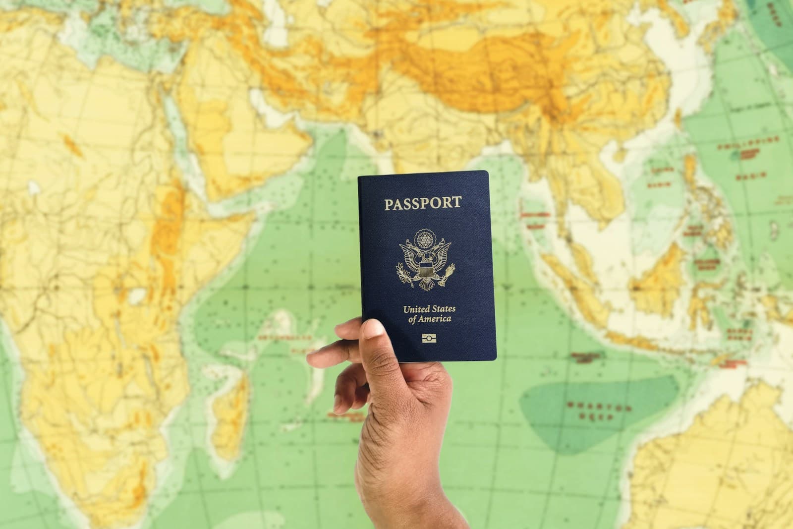 A hand holding a US passport in front of a world map.
